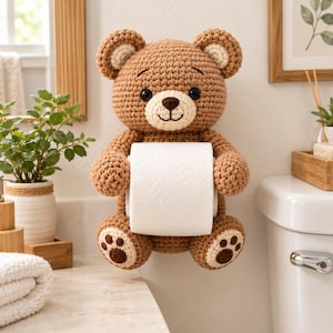 May include: A brown crocheted teddy bear toilet paper holder featuring a cream-colored muzzle, inner ears, and paw pads. The bear has black safety eyes and a stitched dark brown nose and mouth, with its arms positioned to hold a roll of white toilet paper.