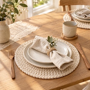 May include: A table setting featuring a round cream-coloured crocheted placemat, two stacked ceramic plates, and a beige linen napkin held by a woven rattan ring. A wooden fork and knife are placed on the sides, with a ceramic mug nearby.