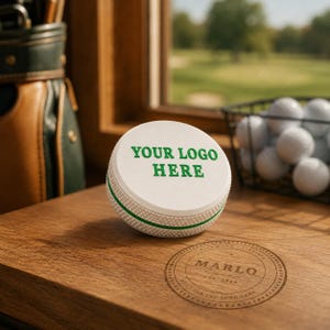 May include: A white circular golf ball marker with a textured side and a green horizontal stripe. The top surface features the green text "YOUR LOGO HERE" in a bold, sans-serif font. The marker sits on a wooden surface with a circular brand logo.