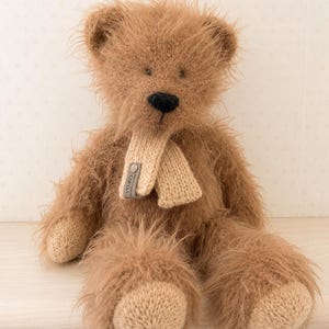 May include: A fuzzy, light brown teddy bear with long, shaggy fur, a dark brown nose, and smooth, light beige knitted paws. The bear wears a matching light beige knitted scarf around its neck, which features a small grey tag labeled YU.KO.