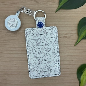 May include: A white rectangular ID badge holder featuring a repeating black line art pattern of gaming controllers, headphones, and the text Game on. The holder includes a blue snap button, a metal split ring, and a retractable white badge reel.