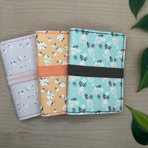 May include: Three rectangular passport holders featuring floral patterns. The left is light purple with white flowers and a matching elastic band. The centre is orange with white flowers and an orange band. The right is teal with white flowers and a dark brown band.