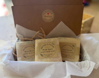 May include: A brown cardboard gift box containing three rectangular bars of goats milk soap wrapped in paper. Each bar features a tan label with the text Heritage & Harvest, identifying the scents as Rosemary, Pink Salt, and Lavender, each 113 grams.
