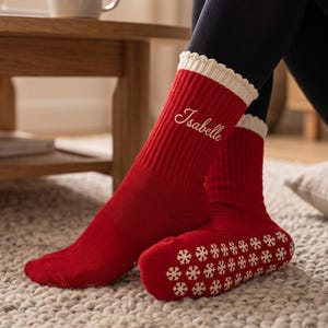 May include: A pair of red ribbed socks with a cream-colored scalloped cuff. The name Isabelle is embroidered in white script on the side of the sock. The sole of the sock features a repeating pattern of white snowflake-shaped non-slip grips.