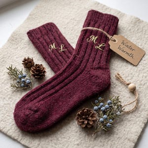 Peut inclure: Une paire de chaussettes en tricot côtelé bordeaux avec les initiales dorées M L brodées sur chaque revers. Une étiquette en papier brun attachée avec de la ficelle porte le texte manuscrit Winter Warmth. Pommes de pin et baies de genièvre.