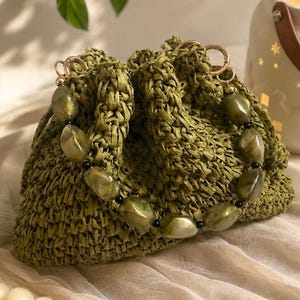 May include: A sage green crocheted drawstring bag featuring a decorative handle made of large, polished, olive-toned marbled beads interspersed with small black spherical beads, attached to the bag with gold-toned metal rings.