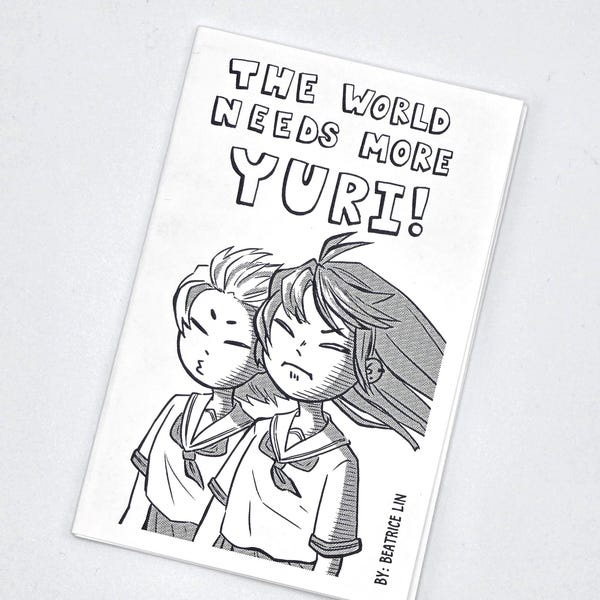 May include: A rectangular white zine with black ink illustrations featuring two characters in school uniforms. The text at the top reads THE WORLD NEEDS MORE YURI! and the bottom right corner includes the text BY: BEATRICE LIN in black ink.