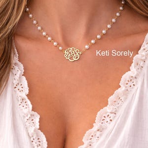Op de afbeelding: Een gouden ketting met een centrale ronde monogramhanger met verstrengelde letters en een ketting bestaande uit kleine witte parels. De tekst Keti Sorely is zichtbaar aan de rechterkant van de afbeelding bij de gouden monogramhanger.