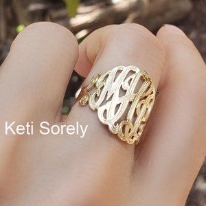 Könnte beinhalten: Ein goldfarbener Ring mit einem Monogramm-Design aus drei ineinander verschlungenen Buchstaben. Der Ring wird an einem Finger getragen.