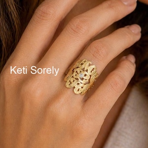 Könnte beinhalten: Ein goldfarbener Ring mit einem aufwendigen, durchbrochenen Design und einem kleinen, klaren Edelstein in der Mitte. Der Ring befindet sich an einem Finger. Der Text "Keti Sorely" ist im Bild zu sehen.