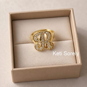 May include: A gold-toned ring featuring a large, cursive letter R as the centerpiece. The letter is encrusted with numerous small, clear, sparkling stones. The ring is presented inside a square, light beige box with the text Keti Sorely visible.