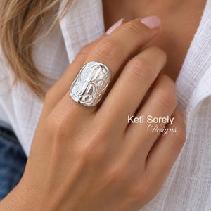 May include: A wide, polished silver ring featuring an intricate, engraved monogram design on its surface. The ring has a smooth, rounded shape that covers a significant portion of the finger. The text Keti Sorely Designs is visible.