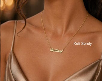 May include: A gold-colored necklace with the name "Brittany" in a cursive font. The necklace is on a delicate chain. The image also includes the text "Keti Sorely". The background is a satin fabric.