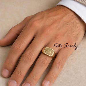 May include: A gold-colored signet ring with the initials "I.J" engraved on the rectangular face. The ring is worn on a finger. The image also includes the text "Keti Sorely".