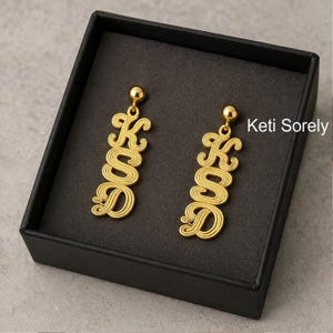 May include: Gold-colored earrings in a black box. Each earring features the letters "KSD" in a cursive, stylized font. The earrings have a small ball at the top for the ear wire. The brand name "Keti Sorely" is visible in the background.