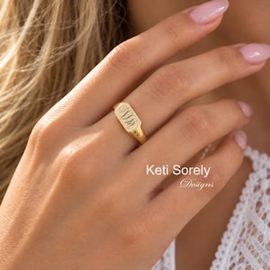 Puede incluir: Un anillo de sello de oro con una cara rectangular alargada y un monograma grabado. La banda de metal pulido tiene un acabado liso y se lleva en un dedo. El texto Keti Sorely Designs es visible en la esquina inferior derecha de la imagen.