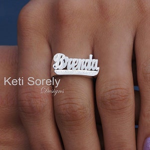 Op de afbeelding: Een zilveren ring met de naam "Brenda" in reliëfletters. De ring heeft een rij kleine, heldere stenen die in de band zijn gezet.