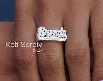 Puede incluir: Un anillo de metal plateado con el nombre Brenda en una fuente cursiva estilizada. La placa con el nombre tiene una superficie texturizada y brillante, montada sobre una banda lisa y pulida. El texto Keti Sorely Designs es visible.