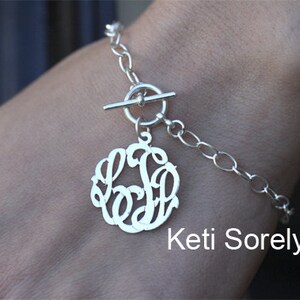 May include: Silver bracelet with a toggle clasp and a monogram charm. The charm is a circle with the letters "K.S." in a script font.