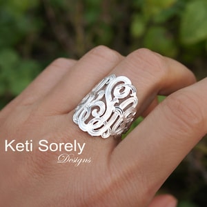 Op de afbeelding: Een zilverkleurige ring met een sierlijk, wervelend monogramontwerp. De ring wordt aan een vinger gedragen en de tekst "Keti Sorely Designs" is zichtbaar in de afbeelding.