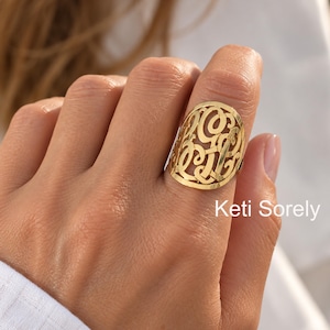 May include: A gold-toned ring featuring an oval-shaped face with an intricate, openwork monogram design. The surface of the ring is polished and reflective. The text Keti Sorely is visible in the lower right corner of the image.