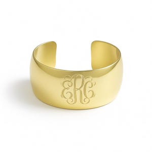May include: A wide, gold-colored cuff bracelet with an engraved monogram. The bracelet has a smooth, polished surface and an open design. The monogram features the letters "ERC" in an elegant, swirling script.