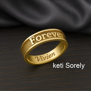 Puede incluir: Un anillo dorado con la palabra "Forever" grabada en la parte superior y el nombre "Vivien" en el lateral. El anillo está sobre un fondo oscuro, con el texto "keti Sorely" en la parte inferior.