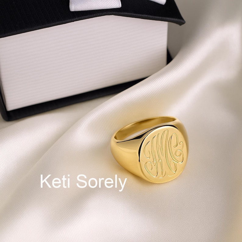 K&ouml;nnte beinhalten: Ein goldener Siegelring mit eingraviertem Monogramm liegt auf einer wei&szlig;en Satin-Oberfl&auml;che. Der Ring hat eine abgerundete Oberfl&auml;che und eine polierte Oberfl&auml;che. Der Text "Keti Sorely" ist sichtbar. Eine schwarz-wei&szlig;e Box befindet sich im Hintergrund.