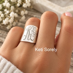 Op de afbeelding: Een brede zilverkleurige ring met een gegraveerd monogram van de letters RC in een decoratief scriptlettertype. Het oppervlak van de ring is glad en gepolijst en reflecteert het licht over de gebogen voorzijde. De tekst Keti Sorely is zichtbaar.