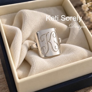 May include: A silver-toned ring featuring a rectangular face engraved with a large, cursive letter K. The ring is presented inside a box with a soft, cream-colored fabric lining. The text Keti Sorely Designs is visible in the upper right corner.