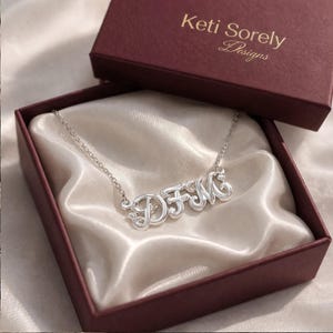 May include: A silver necklace spelling out the letters DFM in a cursive style, presented in an open maroon gift box with a satin lining. The box lid displays the text "Keti Sorely Designs". The necklace is a delicate piece of jewellery.