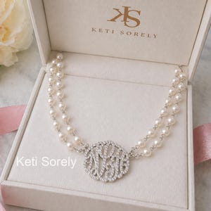Puede incluir: Un collar de perlas con un colgante de monograma, presentado en una caja de joyería blanca. El collar presenta dos hilos de perlas conectados a un colgante circular con un diseño de monograma, rodeado de pequeñas piedras brillantes. La caja lleva el texto "Keti Sorely".