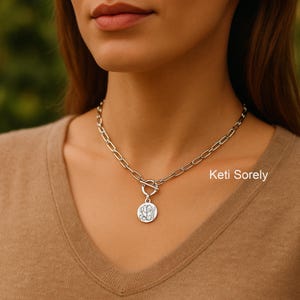 May include: Silver chain necklace with a circular monogram pendant. The necklace has a toggle clasp. The brand name "Keti Sorely" is visible. The necklace is worn on a person.