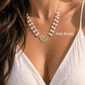 Könnte beinhalten: Eine goldfarbene Halskette mit herzförmigem Anhänger und zwei Perlensträngen. Der Anhänger hat ein aufwendiges Design. Die Halskette wird zu einem weißen Kleid getragen. Der Text "Keti Sorely" ist sichtbar.