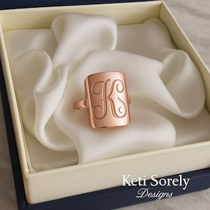 Op de afbeelding: Een roségouden zegelring met de letter "K" gegraveerd op een rechthoekig vlak. De ring wordt gepresenteerd in een met witte satijn gevoerde doos. De tekst "Keti Sorely Designs" is zichtbaar.