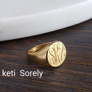 May include: A gold signet ring with an engraved monogram. The ring has a polished finish and a rounded oval face. The text "keti Sorely" is visible in the image.