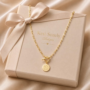 May include: A gold necklace with a circular pendant featuring engraved initials hangs over a beige gift box tied with a satin bow. The box has the text "Keti Sorely Designs" printed on it.