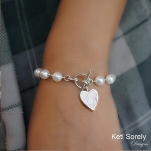 Op de afbeelding: Een zilveren parel armband met een hartvormige bedel gegraveerd met het woord "Hope".