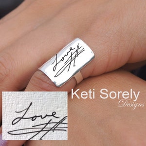 Puede incluir: Un anillo de plata con una banda rectangular grabada con la palabra "Love" y una firma manuscrita. El anillo se lleva en un dedo.