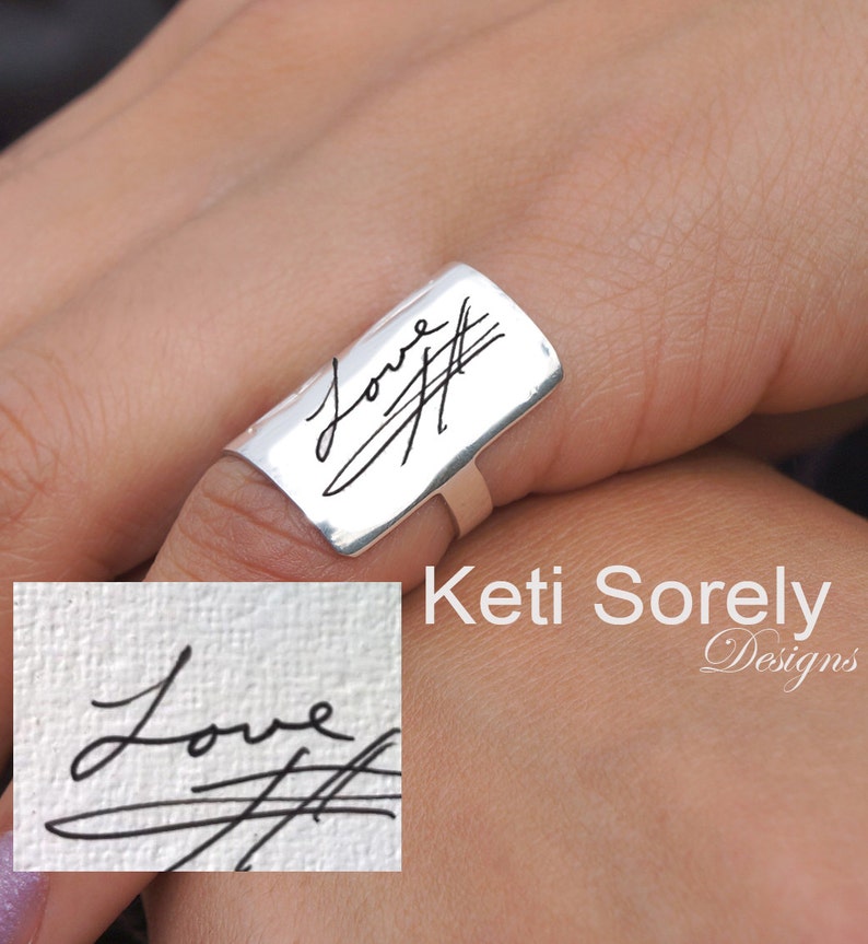 Puede incluir: Un anillo de plata con una banda rectangular grabada con la palabra "Love" y una firma manuscrita. El anillo se lleva en un dedo.