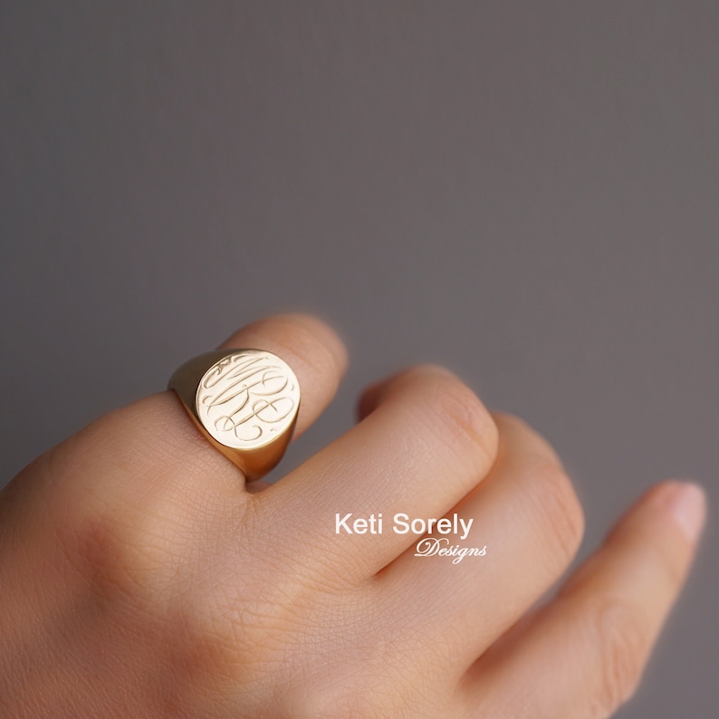 K&ouml;nnte beinhalten: Ein goldener Siegelring mit eingraviertem Monogramm. Der Ring hat eine polierte Oberfl&auml;che und wird am Finger getragen. Das Design des Rings ist auf einer runden Fl&auml;che zentriert. Der Text "Keti Sorely Designs" ist sichtbar.
