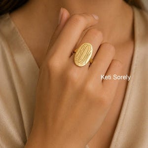 Op de afbeelding: Een gouden ring met een ovaalvormig oppervlak waarop een monogram met de letters BDM in cursief schrift is gegraveerd. De ring heeft een gepolijste afwerking en wordt aan een vinger gedragen. De tekst Keti Sorely is rechtsonder zichtbaar.