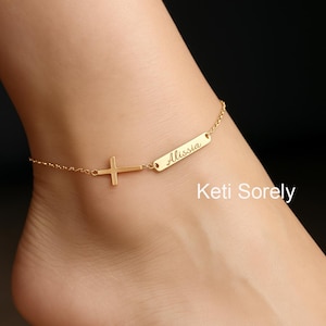 Könnte beinhalten: Goldfarbenes Fußkettchen mit einem Kreuzanhänger und einem rechteckigen Schild mit dem eingravierten Namen "Alissia". Die feine Kette verbindet das Schild und das Kreuz. Der Text "Keti Sorely" ist sichtbar.