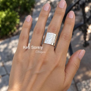 May include: A wide silver band ring featuring an engraved script monogram on the front surface. The metal has a polished finish and the ring is worn on the middle finger. The text Keti Sorely Designs is visible on the image.