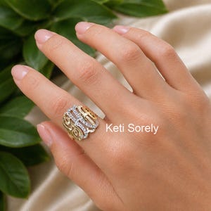 Puede incluir: Un anillo de oro y plata con un diseño decorativo con piedras preciosas transparentes. El anillo está en el dedo de una persona. El texto "Keti Sorely" es visible en la imagen.