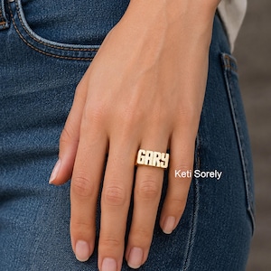 May include: A gold-toned ring featuring the name GARY in bold, block capital letters. The ring has a polished, metallic finish and is worn on a finger. The text Keti Sorely is visible in white lettering on the right side of the image.