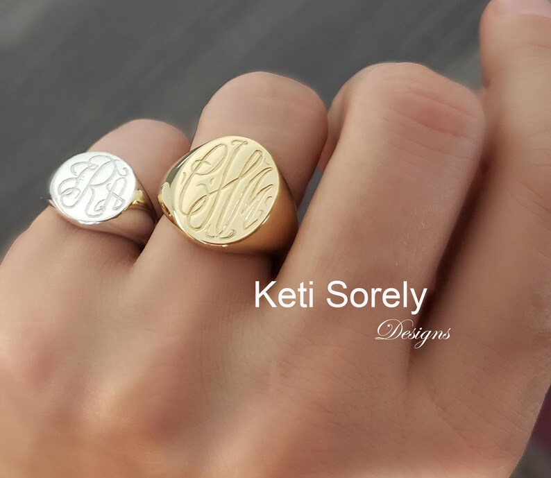 K&ouml;nnte beinhalten: Zwei gravierte Siegelringe, einer aus Gold und einer aus Silber, die jeweils elegante Monogramm-Initialen zeigen. Der goldene Ring ist gr&ouml;&szlig;er und auff&auml;lliger, w&auml;hrend der silberne Ring kleiner ist und einen wei&szlig;en Hintergrund hat. Die Ringe werden an einer Hand getragen.