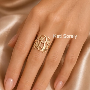 May include: A gold monogram ring with the letters "B" and "R" in a decorative script. The ring is displayed on a finger, highlighting its detailed design. The text "Keti Sorely" is visible in the background.