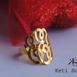 May include: A gold-colored ring with a delicate, intricate design featuring a monogram. The ring is on a white surface with a red fabric in the background.