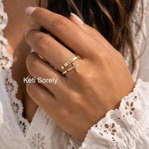 May include: A gold ring featuring a cross pendant and the name "Vivvi" engraved on the band. The ring is displayed on a finger. The image also shows the text "Keti Sorely".
